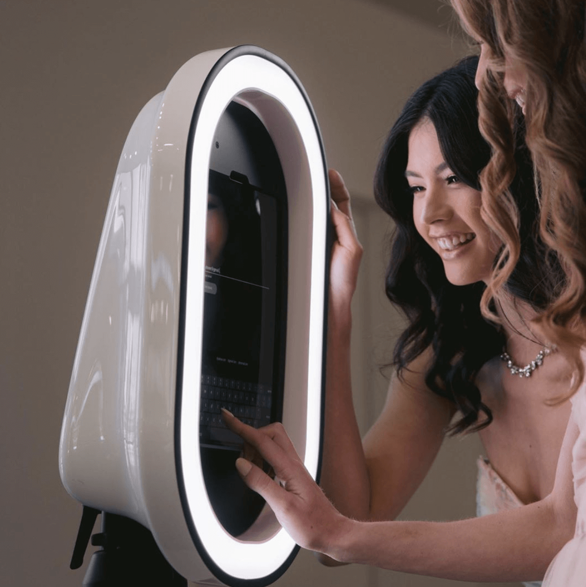 two women smiling and touching a photo booth with a ring light in the shape of an oval.