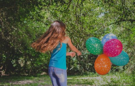 A girl is running through a wooded area with colorful balloons.