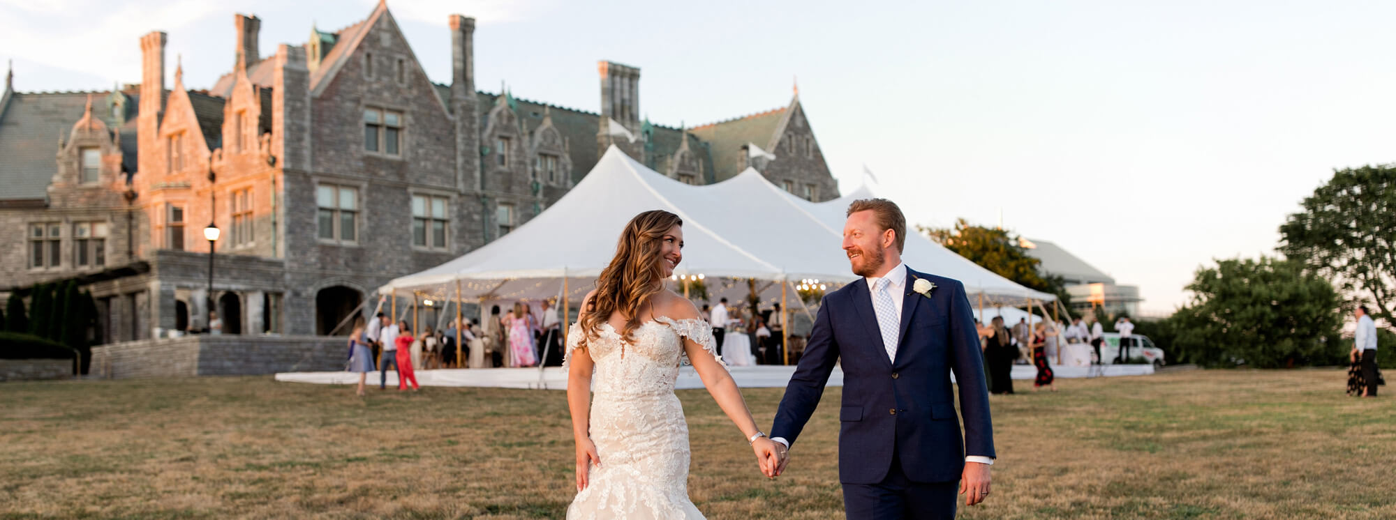 A bride and groom hold hands and walk on grass in front of a large stone building—one of the most picturesque wedding venues in CT—and a white event tent filled with guests.
