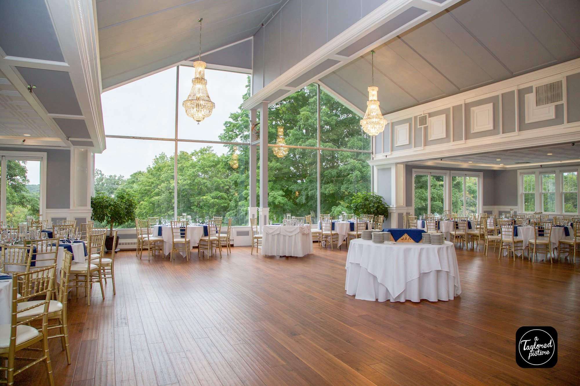 Elegant banquet hall with large windows, chandeliers, wooden floor, and round tables set for an event. Perfect for couples seeking wedding venues in CT, with beautiful trees visible just outside the windows.
