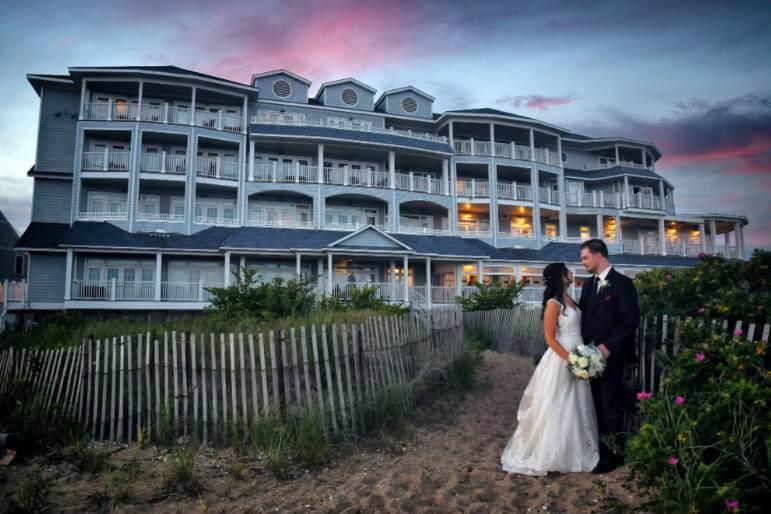 A bride and groom stand together on a sandy path with a bouquet, in front of one of the most stunning wedding venues in CT—a large, multi-story beachfront hotel at sunset.