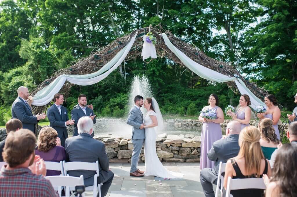 A couple kisses at an outdoor wedding ceremony under a decorative arch with draped fabric. Surrounded by the wedding party and guests, they celebrate their love at one of the charming wedding venues in CT, with a fountain and lush greenery creating a picturesque backdrop.