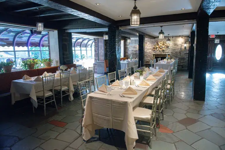 This elegant banquet room, perfect for wedding venues in CT, features long tables set with white tablecloths, folded napkins, and floral centerpieces. Stone walls and a cozy fireplace add to the charming atmosphere.