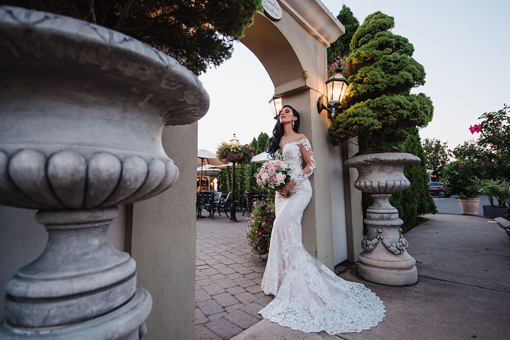 A bride in a white lace wedding dress holding a bouquet stands under an archway outdoors, surrounded by greenery and large planters at one of the most beautiful wedding venues in CT.