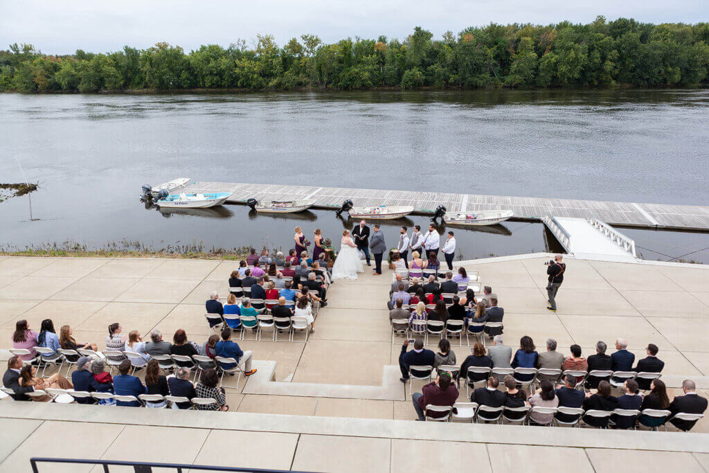 Outdoor wedding ceremony beside a river at one of the scenic wedding venues in CT, with guests seated in rows, a wedding party at the front, and boats docked along a wooden pier in the background.
