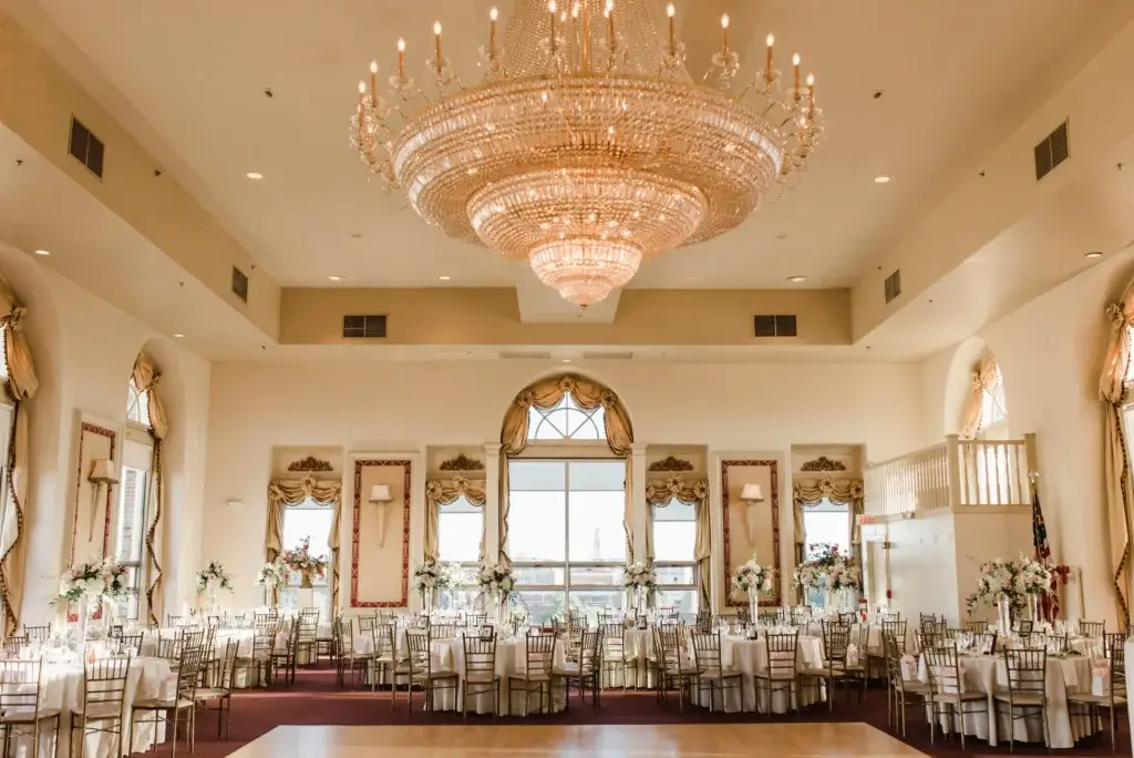 A spacious, elegant banquet hall—perfect for wedding venues in CT—features a large crystal chandelier, decorated tables with white linens and floral centerpieces, and tall windows letting in natural light.