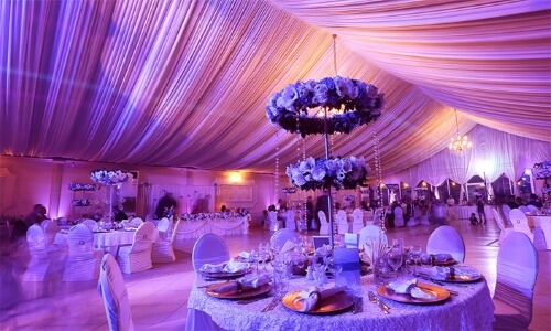 Wedding venue with ceilings draped in white material lit up with purple uplighting. There is a large floral chandelier and tables set with white linens and tableware.