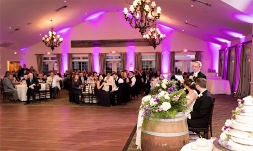Wedding venue filled with guests sitting at tables. The room is lit with bright purple uplighting and the bride and groom sit on the right side of the picture at their bridal table.