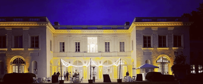 An illuminated, large two-story building at night with people gathered outside, featuring arched windows and a central balcony, showcasing expert lighting services.