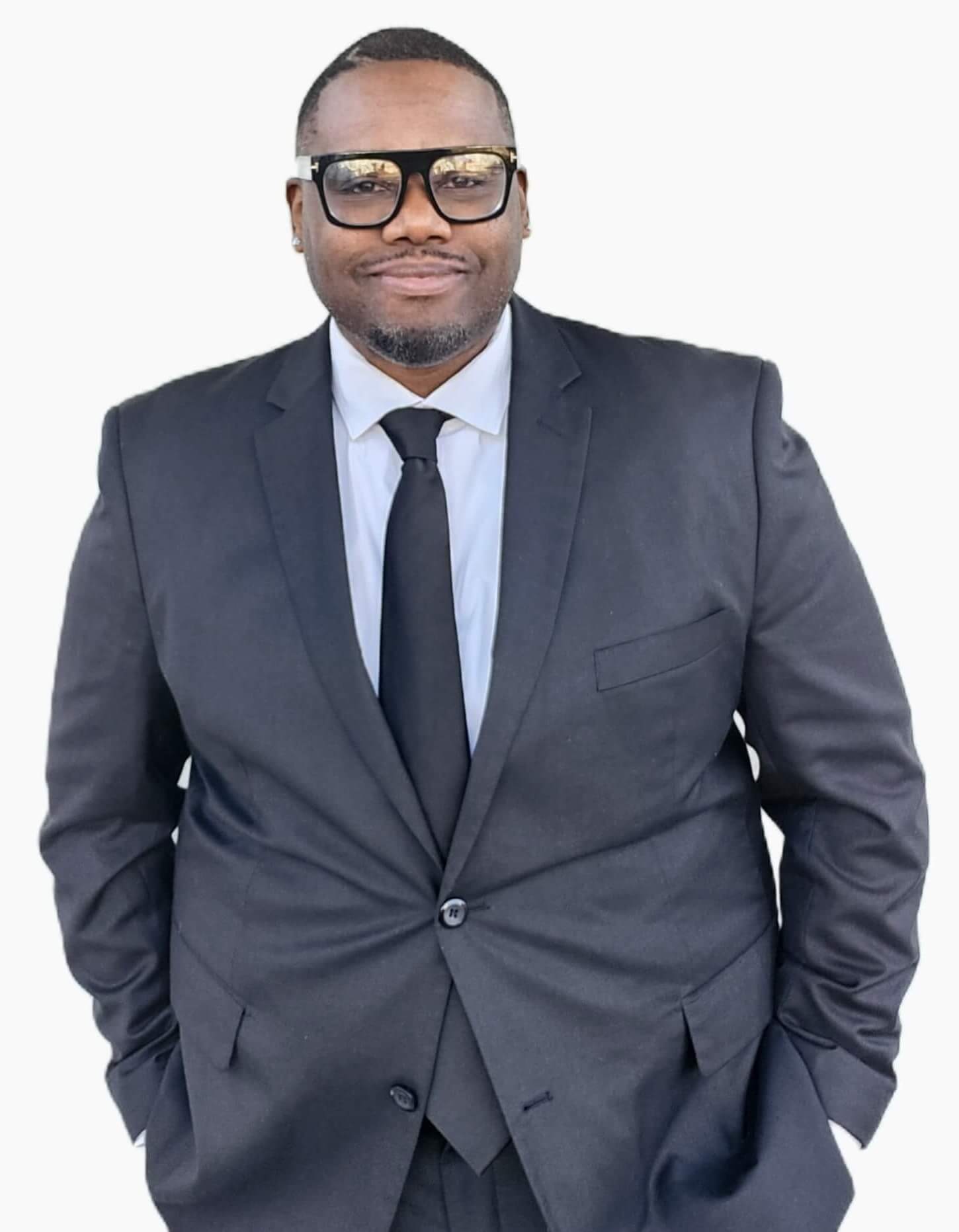 DJ-Maurice5 A man wearing a black suit, white shirt, and black tie stands with his hands in his pockets against a plain white background.