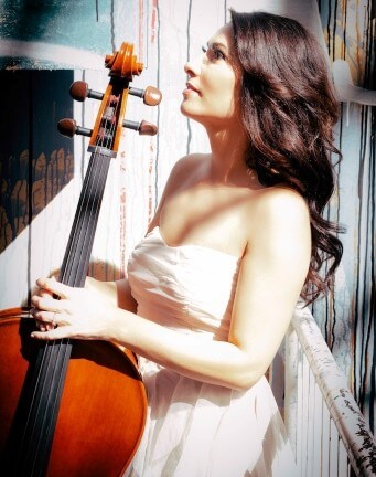 A woman in a white strapless dress holds a cello and looks upward. Sunlight highlights her face and the instrument, with a painted wooden backdrop behind her.
