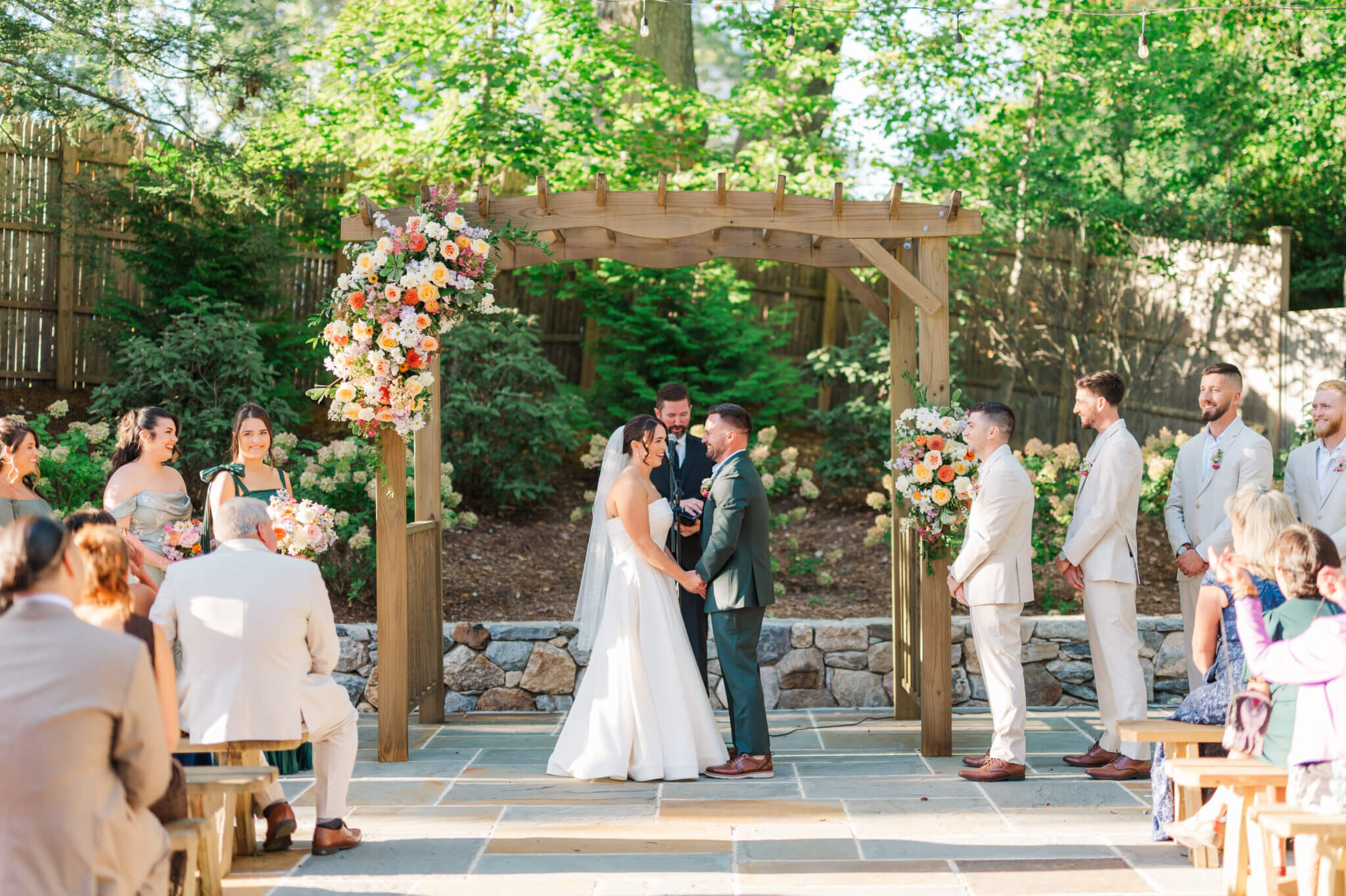 A bride and groom stand facing each other at an outdoor wedding ceremony under a wooden arch, surrounded by their wedding party and seated guests at the Hops Company in CT