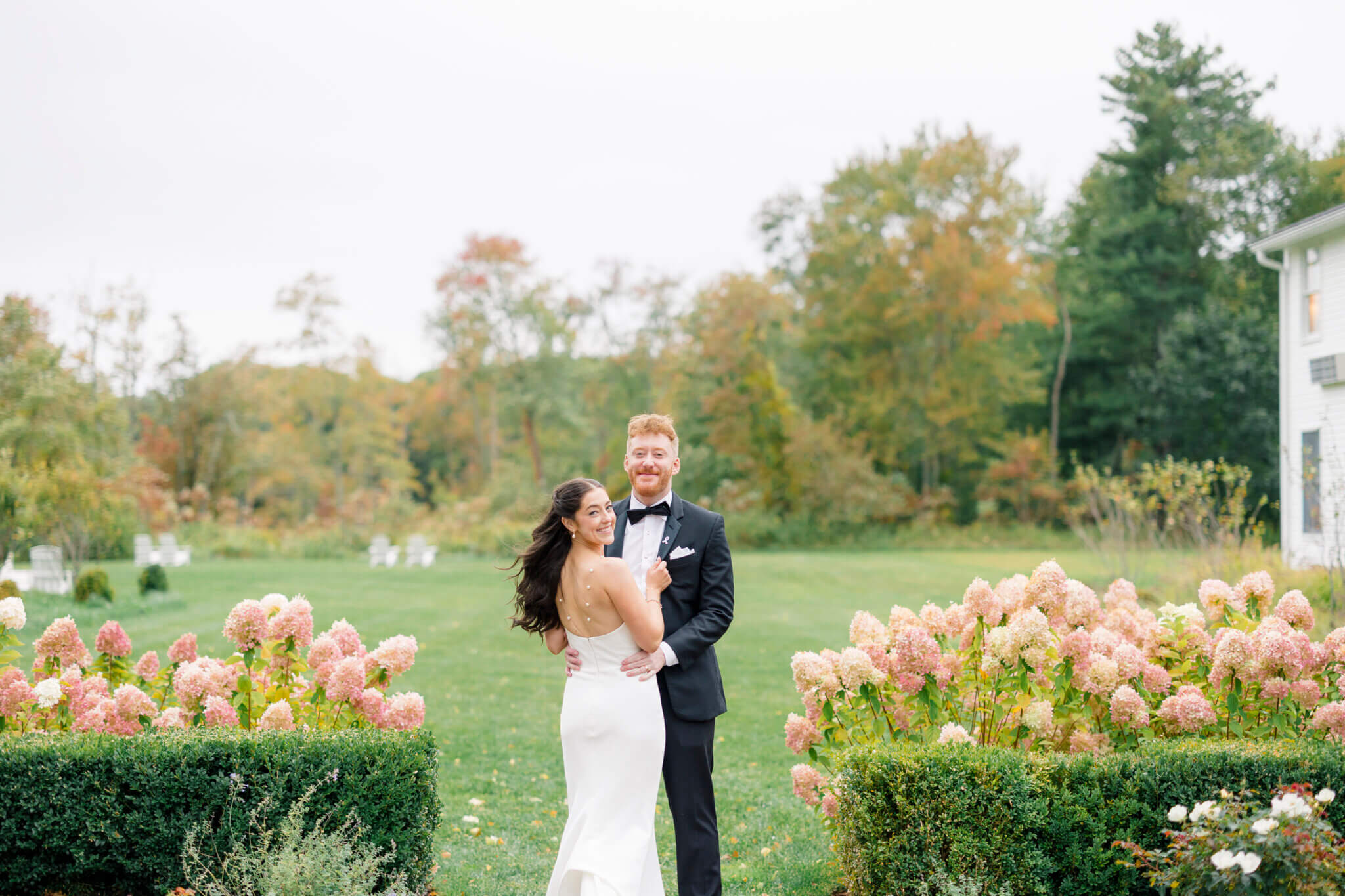 A bride and groom stand together outdoors in formal attire at one of the scenic wedding venues in CT, surrounded by blooming bushes, greenery, trees, and a charming house.