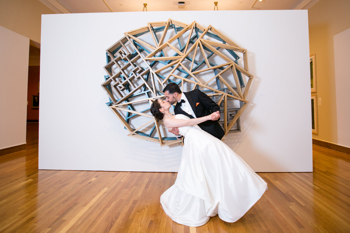 A bride and groom in formal attire share a dip kiss in front of a geometric wall sculpture on a hardwood floor, capturing the modern elegance found at top wedding venues in CT art galleries.