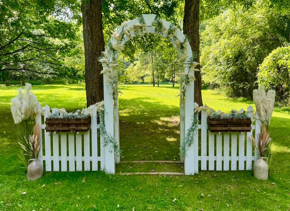 A white picket fence and arch decorated with greenery and pampas grass stands on a grassy area between two trees in a sunlit park, offering charming inspiration for wedding venues in CT.