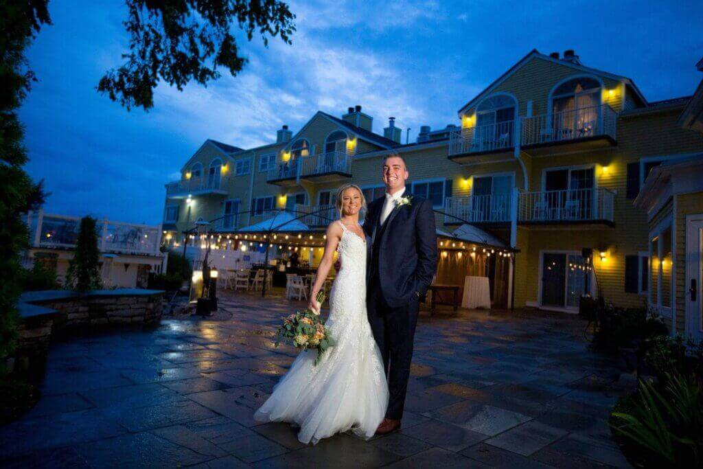 A newlywed couple poses outdoors in wedding attire at dusk in front of a well-lit building with balconies, showcasing one of the most elegant wedding venues in CT.