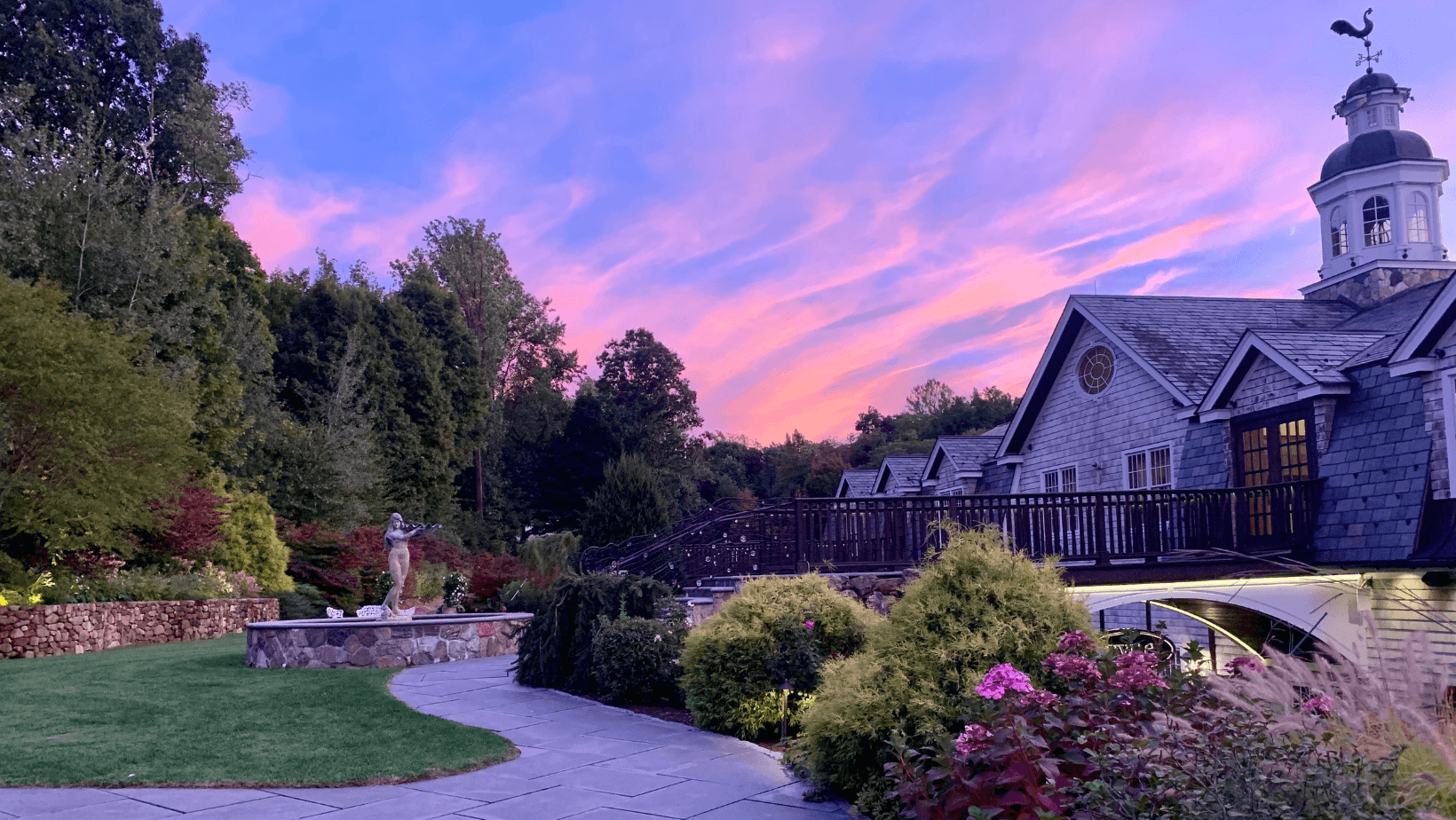 Landscaped garden with flowers, a stone path, and a statue in front of a large house at sunset—an ideal setting among top wedding venues in CT, with trees and a colorful sky painting the perfect backdrop.